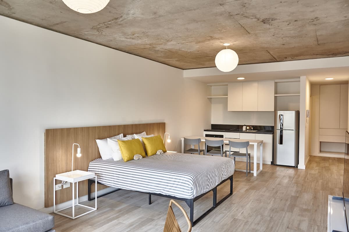 A modern, well-lit bedroom with a bed, bedside table, and a small seating area. Adjacent is a kitchenette with a refrigerator and a dining table.