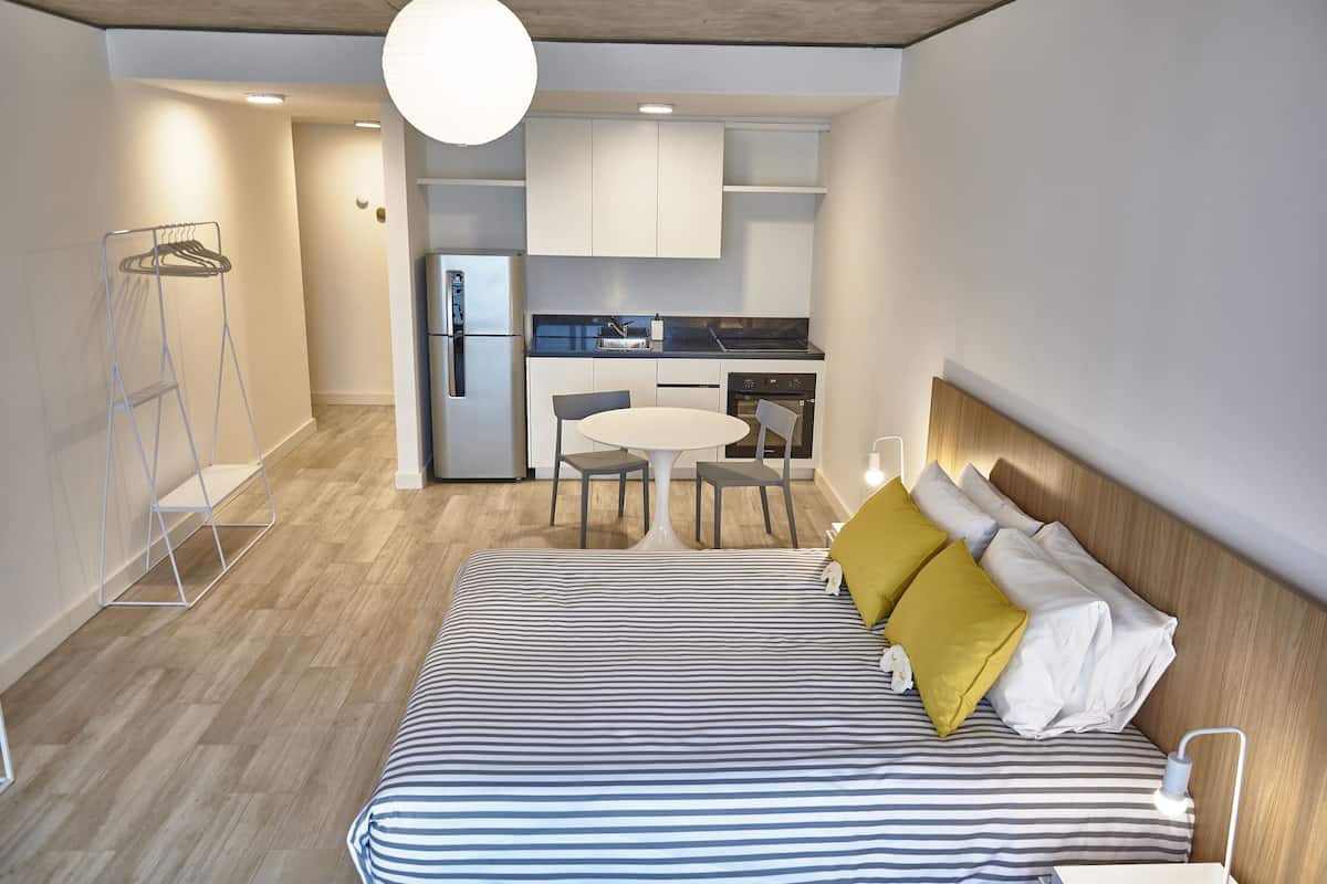 A modern hotel room with a bed, a small dining table, and a kitchenette.