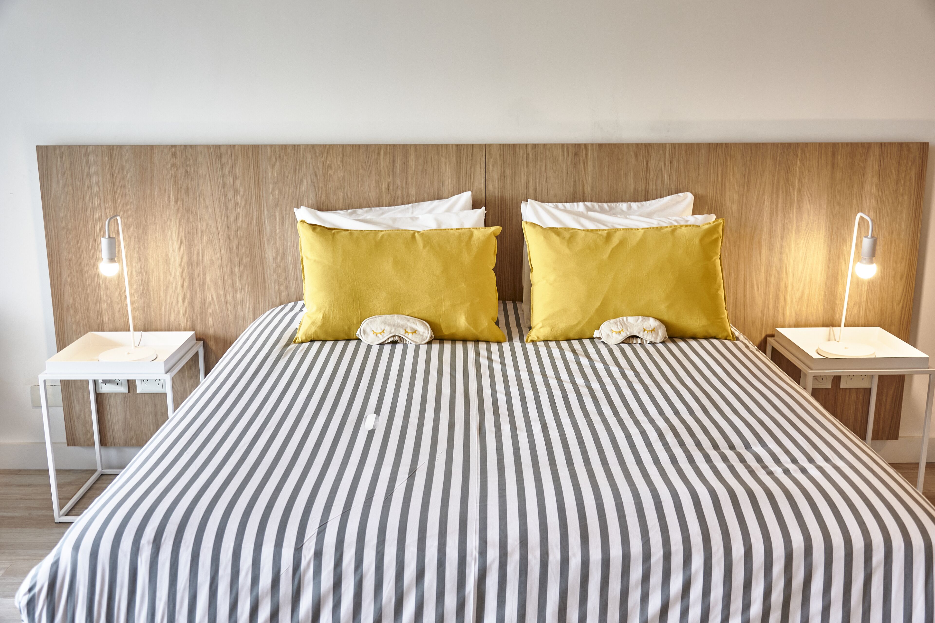 Een bed met gestreepte lakens, twee gele kussens en twee nachtkastjes met lampen.
