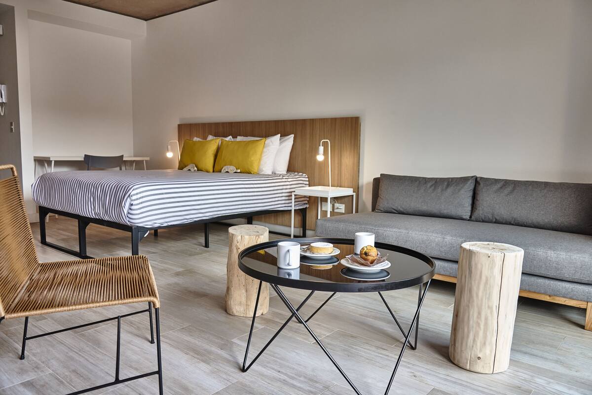 A modern hotel room with a bed, a sofa, a dining table, and a wicker chair.