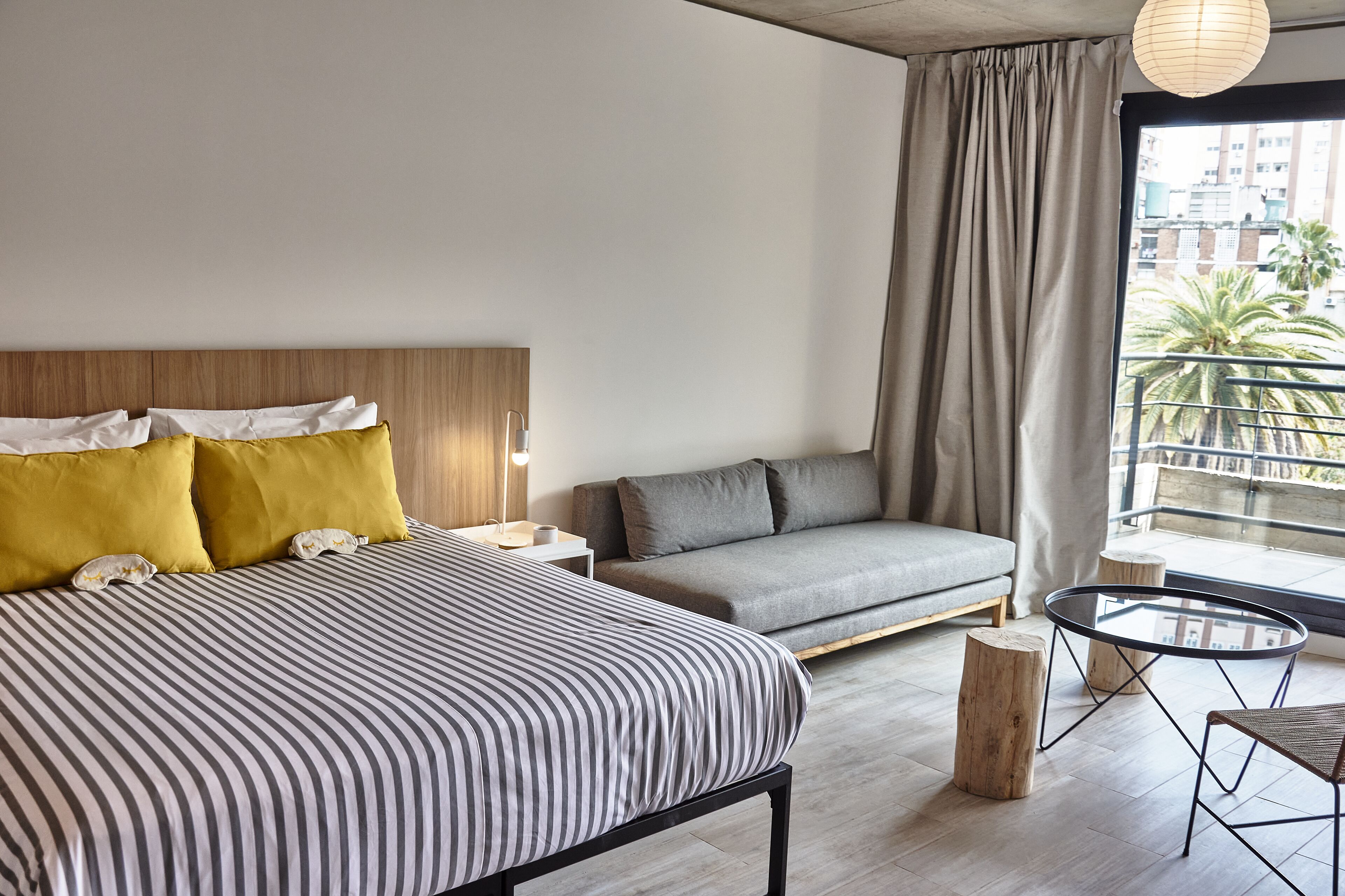 A modern hotel room with a large bed, a sofa, a small table, and a view of the city through a large window.