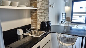 Een moderne keuken met een zwart aanrecht, een roestvrijstalen spoelbak en een accentmuur van baksteen. Er zijn witte kastjes, een koffiezetapparaat en een stoel bij het raam.