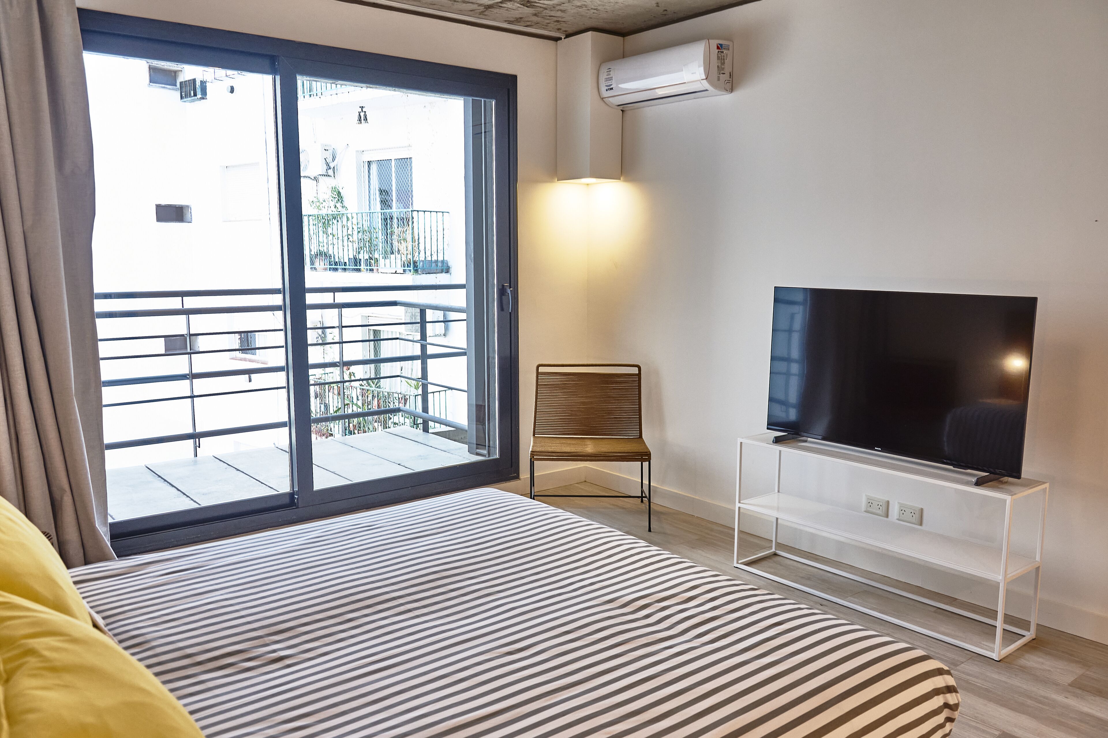 A modern bedroom with a large bed, a TV on a white stand, and a balcony with a railing.