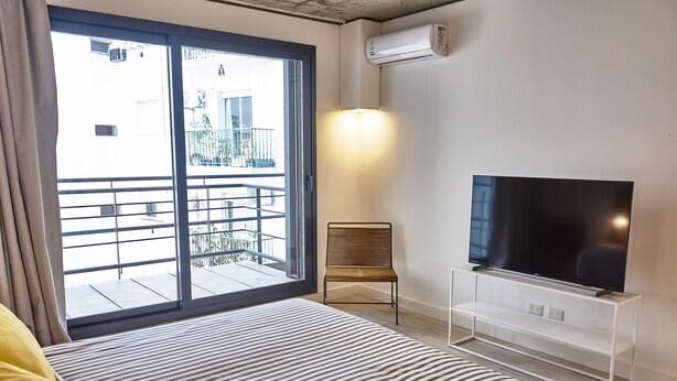 A modern bedroom with a large bed, a TV on a white stand, and a balcony with a railing. - Babel Botánico (Buenos Aires)