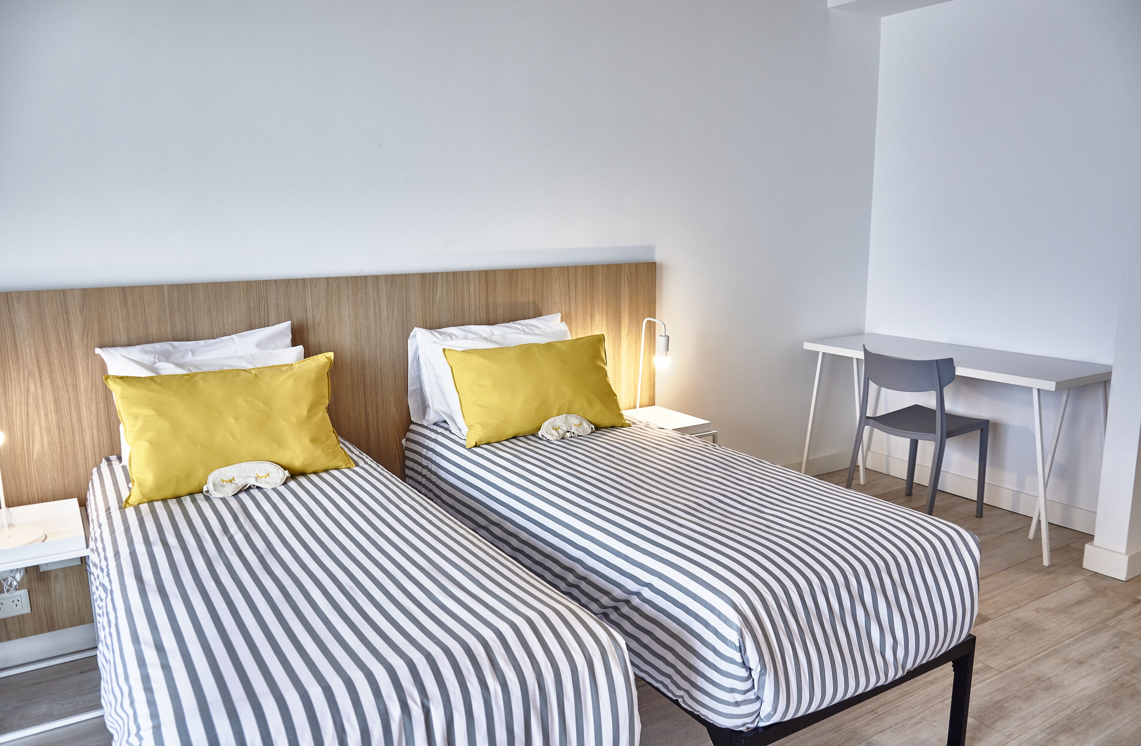 Une chambre d’hôtel avec deux lits, chacun doté de draps à rayures et d’un oreiller jaune. On y trouve un bureau avec une chaise et une table de chevet avec une lampe.