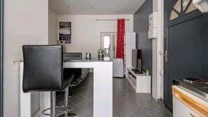 En kompakt opholdsstue med tekøkken, spiseplads med en sort stol, et lille køleskab og et fjernsyn.