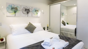 Un lit bien fait, avec du linge de lit blanc, une couverture grise et deux serviettes pliées posées sur le lit. Trois œuvres d’art, accrochées au mur, représentent des motifs floraux abstraits. Une petite table avec une bouteille et un verre se trouve à côté du lit.