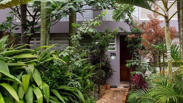 Un jardin luxuriant, avec un chemin de pierre menant à l’entrée d’un bâtiment, dont la porte est marquée du numéro 29A.