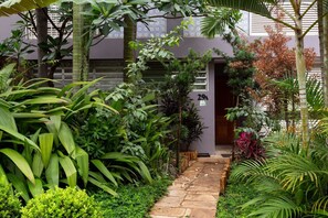 Un jardin luxuriant, avec un chemin de pierre menant à l’entrée d’un bâtiment, dont la porte est marquée du numéro 29A.