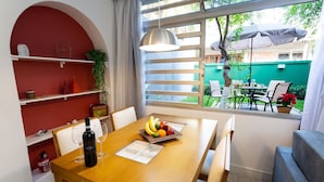 Un coin repas avec une table en bois, des chaises, un panier de fruits, des verres à vin et une bouteille. Il y a une fenêtre donnant sur l’extérieur, un luminaire suspendu et un mur rouge avec des étagères.