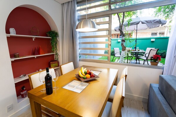 Ein Essbereich mit Holztisch, Stühlen, einer Obstschale, Weingläsern und einer Flasche. Ein Fenster bietet Blick ins Freie, eine Pendelleuchte und eine rote Wand mit Regalen.