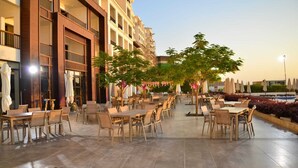 An outdoor cafe area with tables and chairs, surrounded by a building with balconies and a pool in the background. - PORTO & ALMIRA BEACH (Sweimeh)