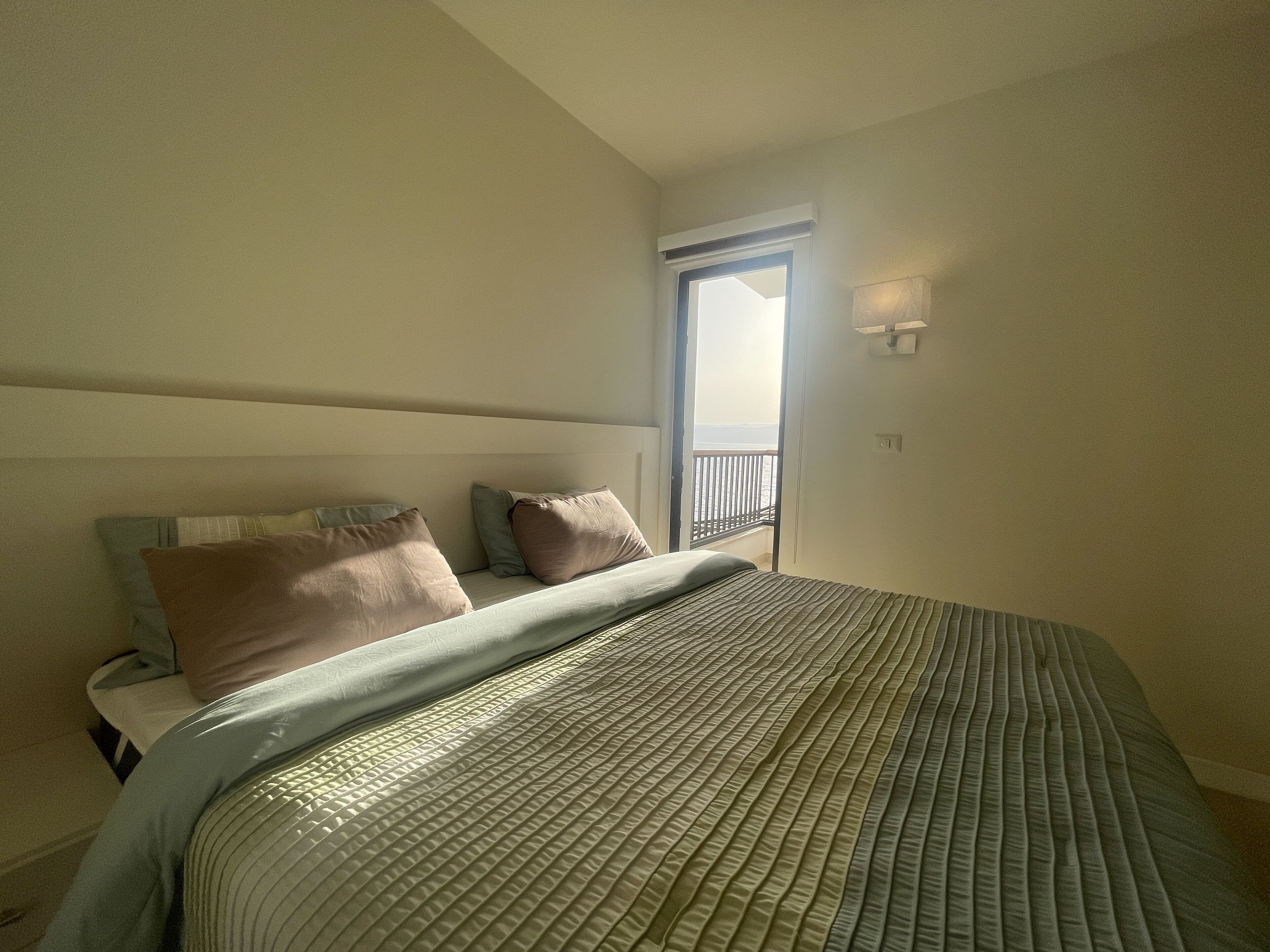 Quarto com cama, almofadas, mesa de cabeceira, janela e varanda.