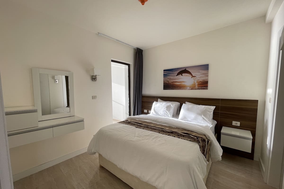 A hotel room with a large bed, a nightstand, a dresser, a mirror, and a painting of a dolphin jumping out of the water.