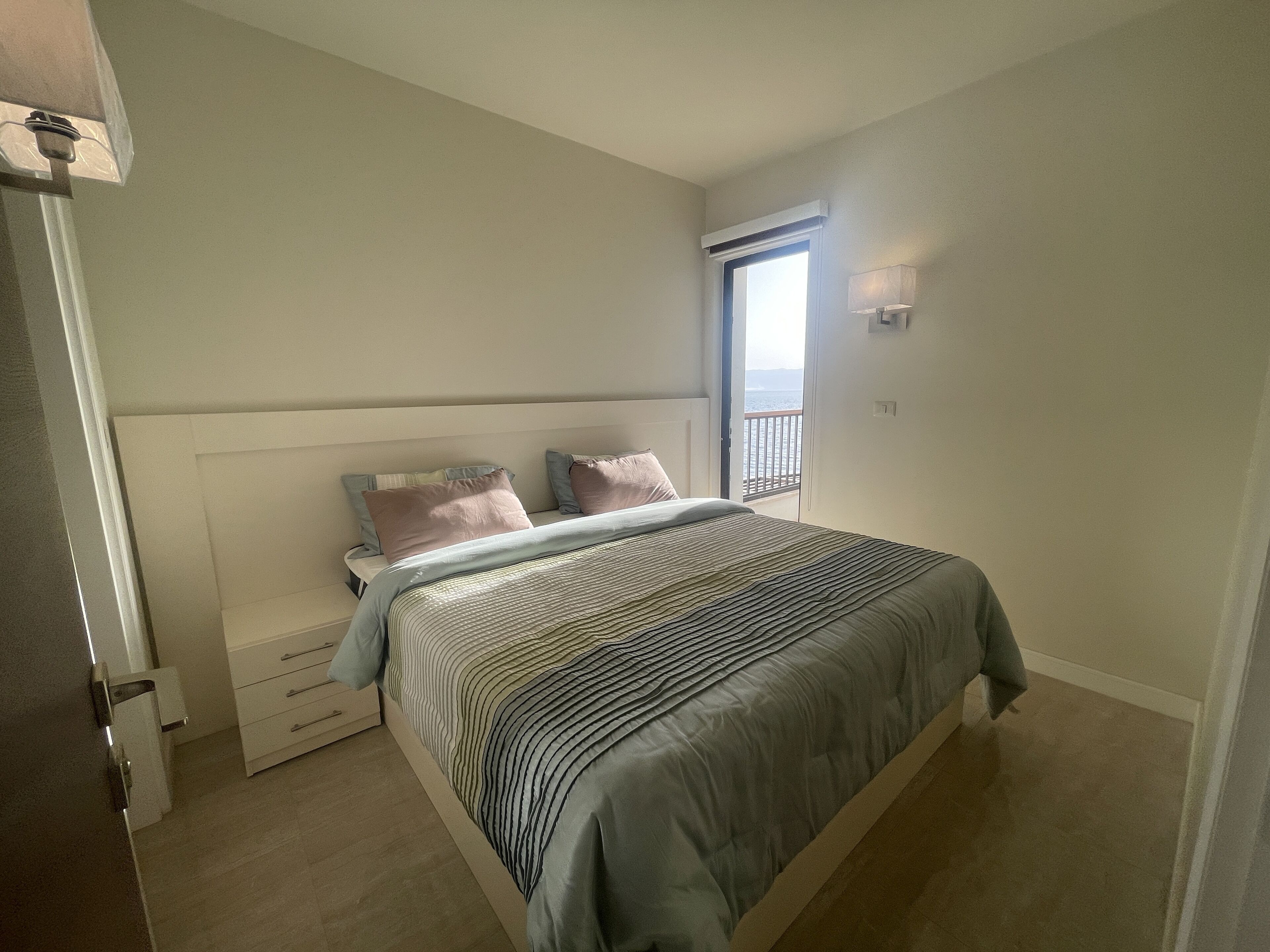 A bedroom with a bed, bedside tables, a window, and a view of the sea.