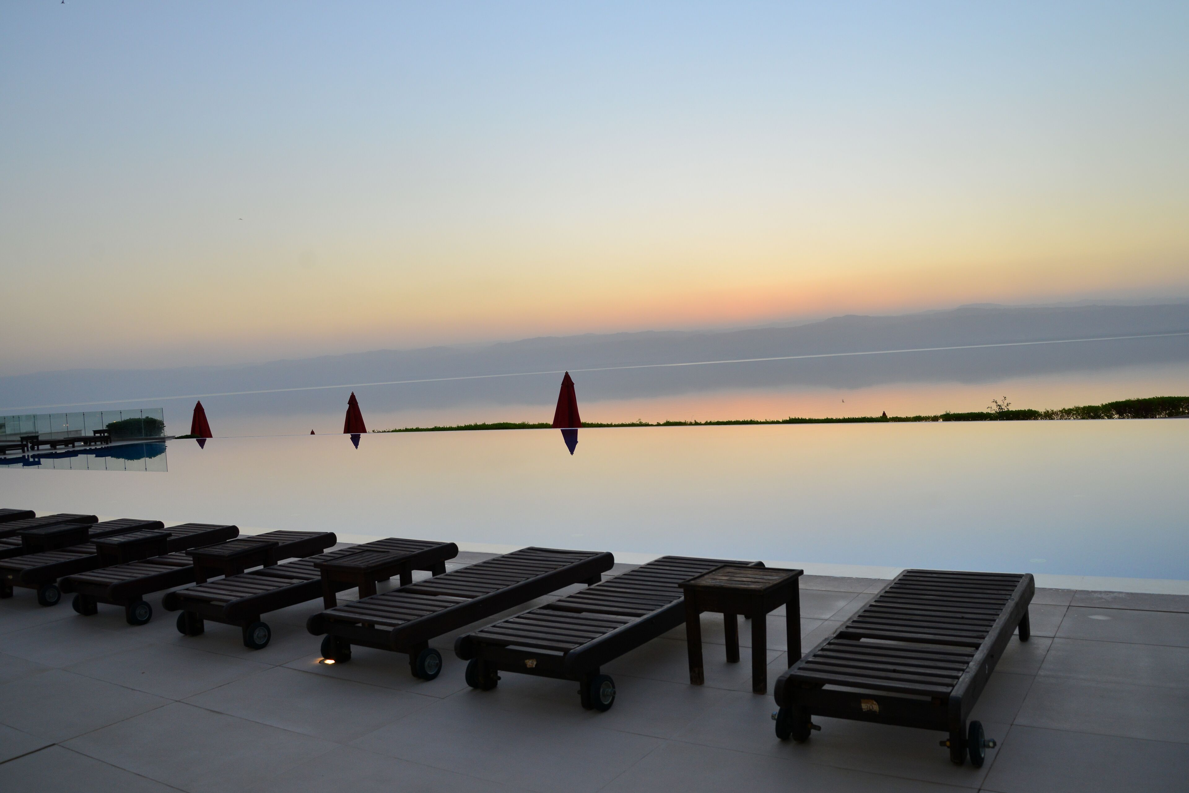Un tranquilo espacio lounge frente al agua, con varias sillas y mesas de madera, un cuerpo de agua en calma y un cielo despejado.