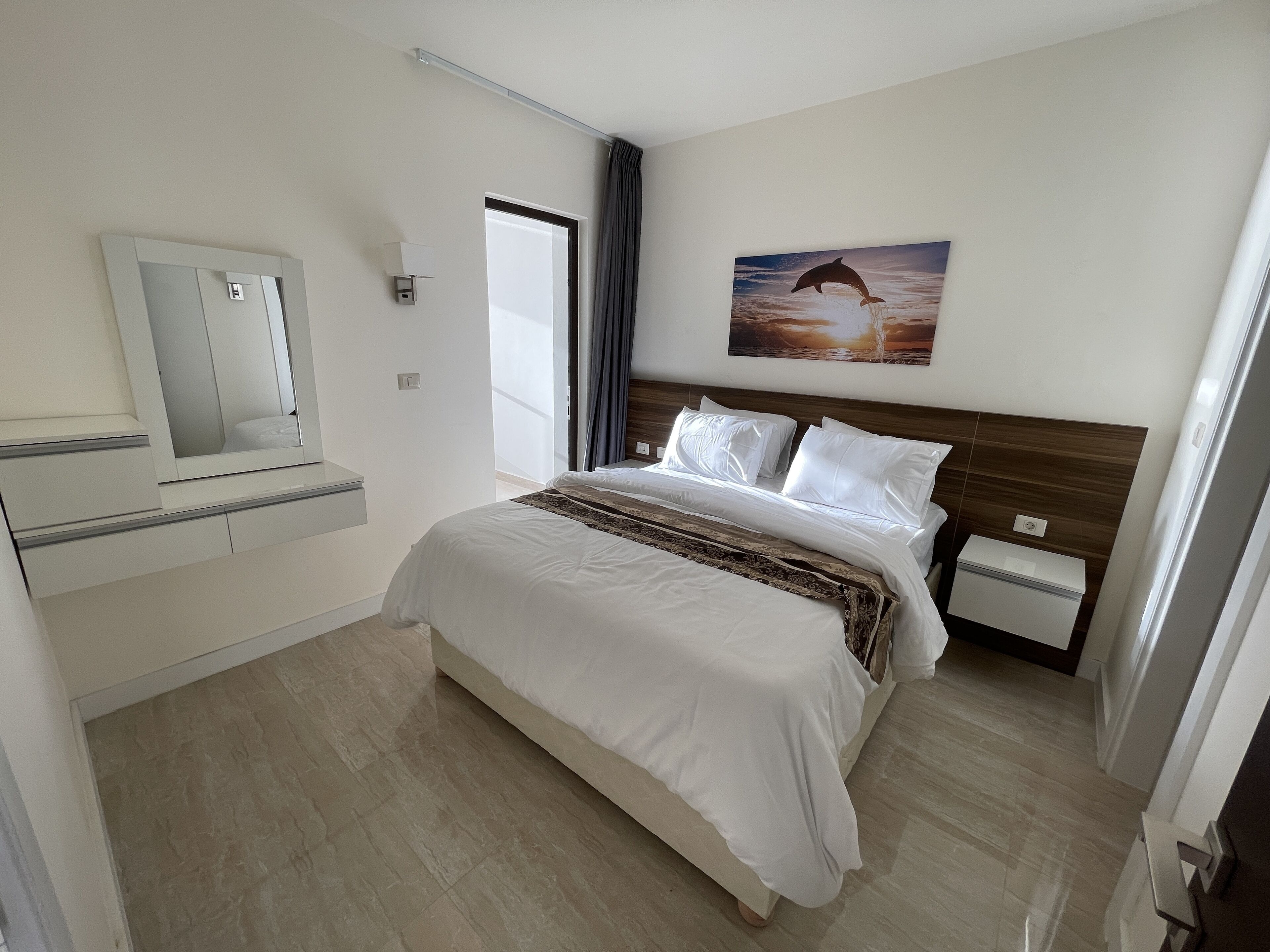 A hotel room with a bed, a nightstand, a bedside table, a mirror, a window with curtains, and a wall-mounted picture.