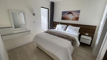 A hotel room with a bed, a nightstand, a bedside table, a mirror, a window with curtains, and a wall-mounted picture.
