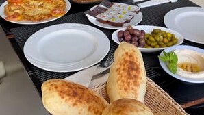 A dining table set with various dishes, including a pizza, salad, and bread, in a modern dining area with glass partitions and cushioned chairs. - PORTO & ALMIRA BEACH (Sweimeh)