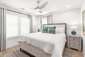 A bedroom with a bed, nightstands, a ceiling fan, and a window with curtains. - Sweet Retreat by Tybee Vacation Rentals (Tybee Island)