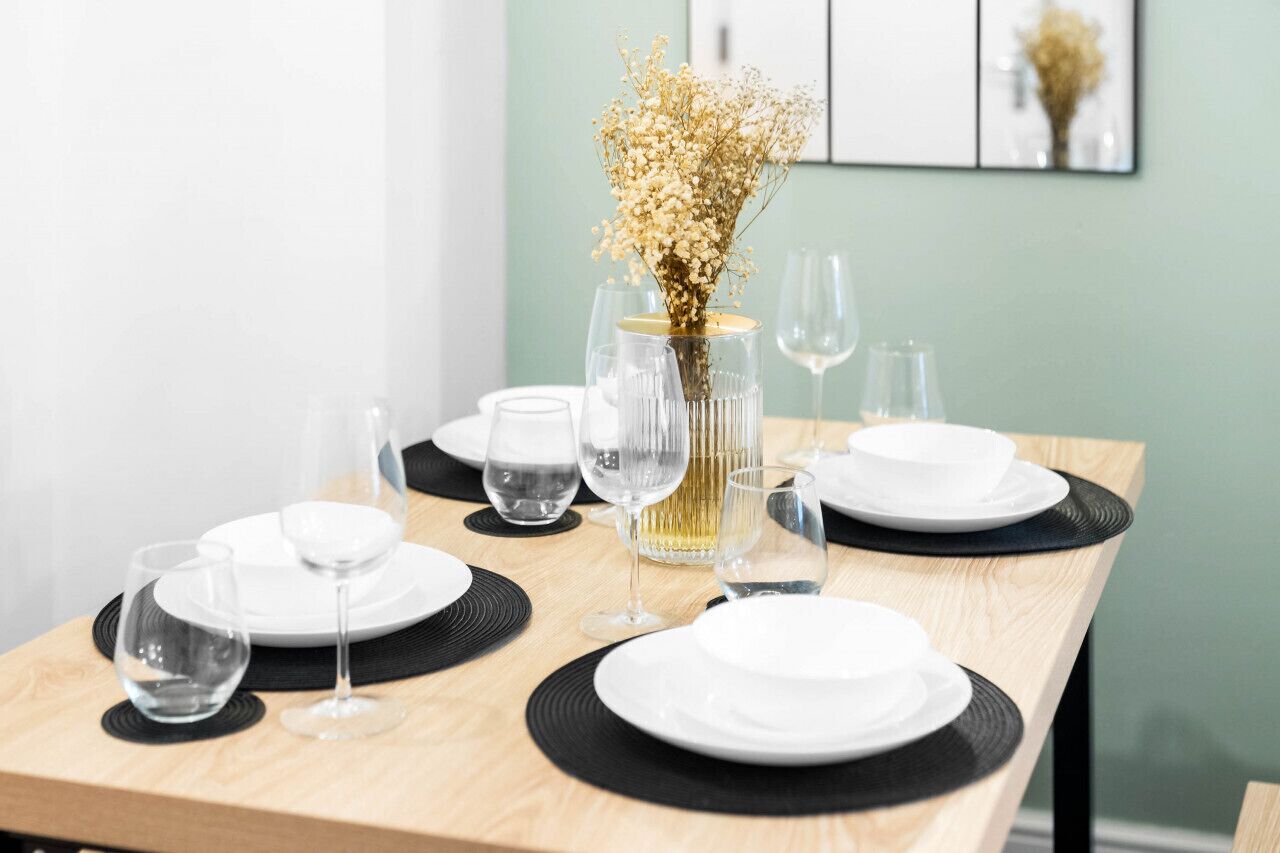 Une table à manger est dressée avec de l’assiette blanche, des sets de table noirs et des verres à vin transparents. Un vase contenant des fleurs séchées est placé au centre.
