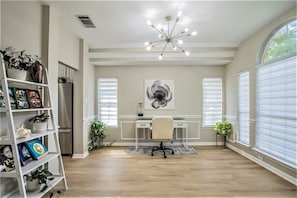 Um quarto amplo com piso de madeira, uma escrivaninha com cadeira, uma estante de livros, uma janela grande com persianas e um lustre.