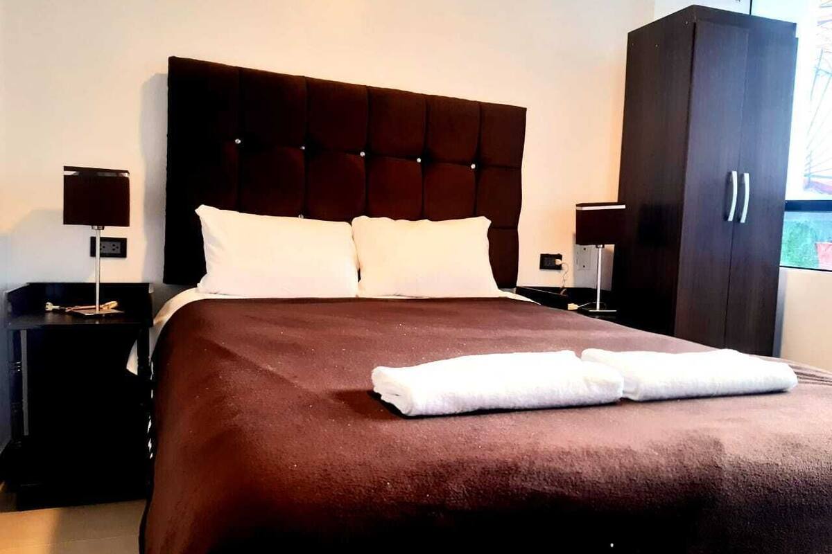 Un lit bien fait, recouvert d’une couverture marron et agrémenté d’oreillers blancs, est flanqué de deux tables de chevet sombres sur lesquelles reposent des lampes, dans une pièce où se trouve une grande armoire.