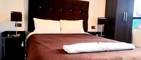 A neatly made bed with a brown bedspread and white pillows, flanked by dark nightstands with lamps, in a room with a large wardrobe.