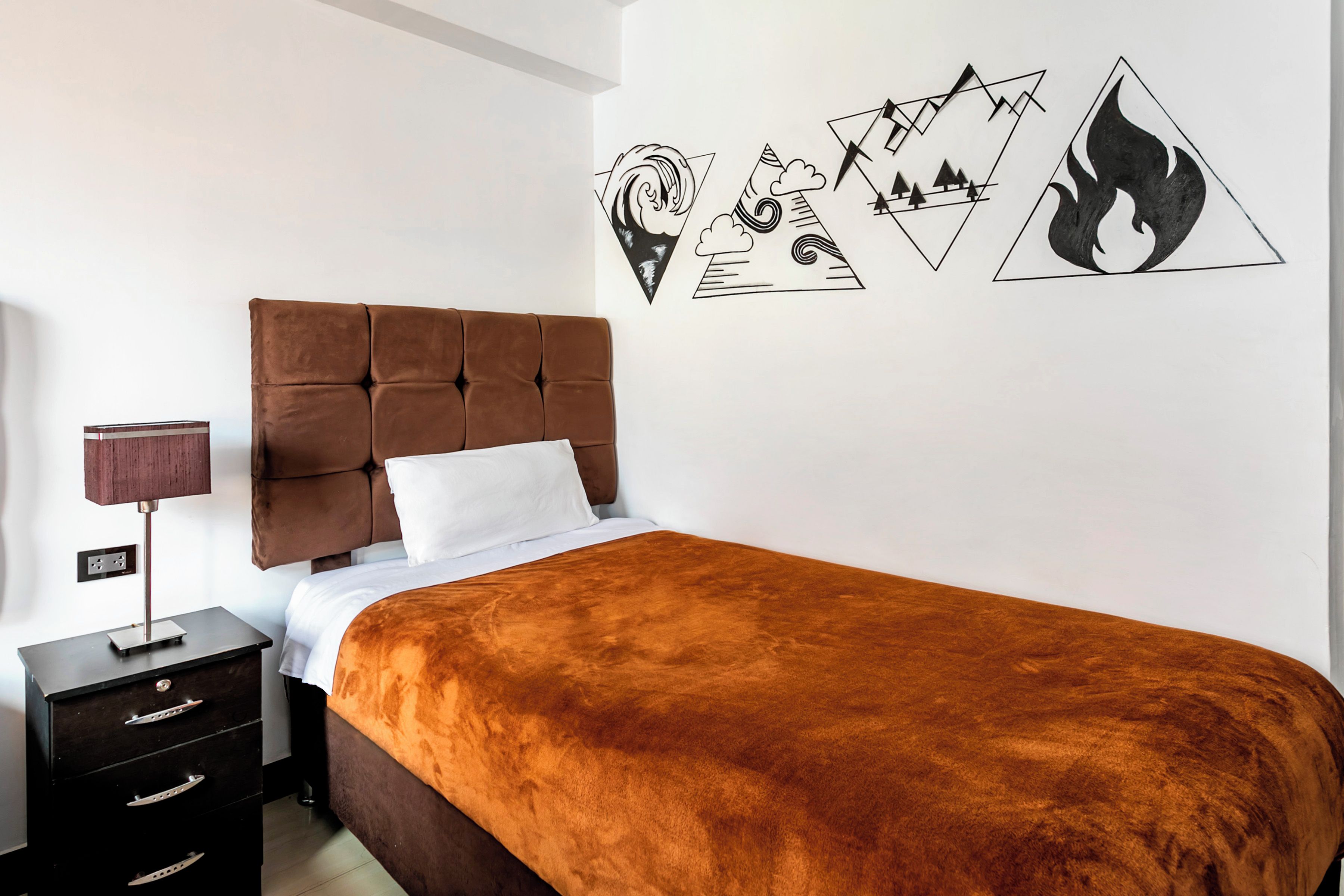 A bedroom with a brown upholstered headboard, a white pillow, a brown blanket, a bedside table with a lamp, and a wall with geometric wall art.