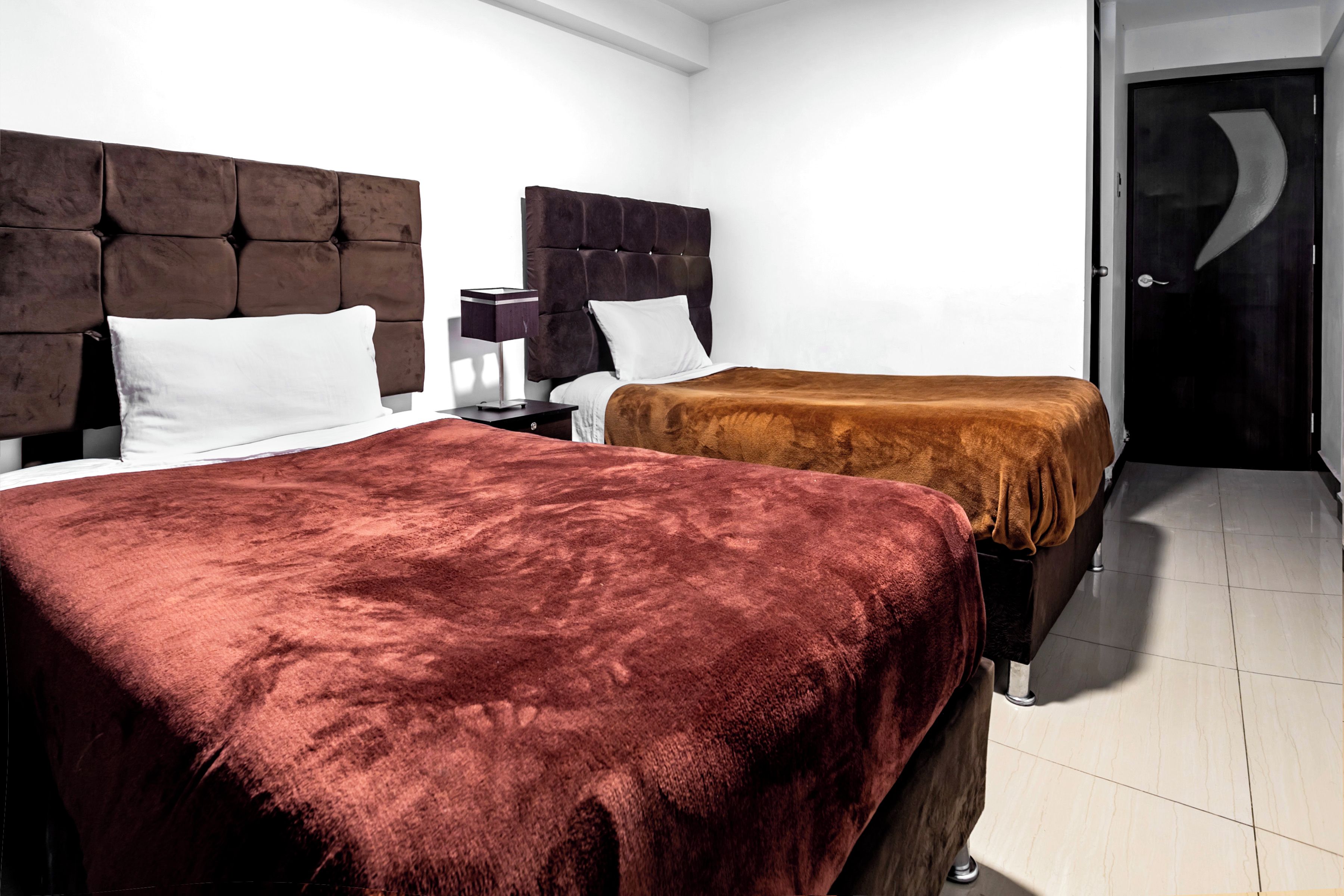 Quarto de hotel com duas camas, uma mesa de cabeceira, uma luminária e uma porta.