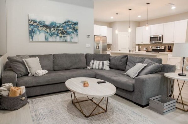 A modern living room with a grey sectional sofa, a round coffee table, and a kitchen in the background. - New, charming 3-bedroom home with pool (Nokomis)