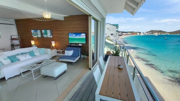 Ein modernes Wohnzimmer mit einer Couch, einem Fernseher und einem Balkon mit Blick auf einen Strand.