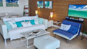 Ein modernes Wohnzimmer mit einem weißen Sofa, einem blauen Sofa, einem Marmortisch und einem weißen Hocker. An der Wand ist ein Fernseher angebracht, ein kleiner Tisch mit einer Lampe steht daneben, und ein Regal zeigt dekorative Gegenstände.