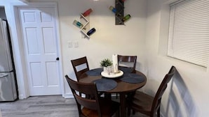 Uma área de refeições com uma mesa redonda, cadeiras, um lustre pendurado e uma prateleira na parede com objetos coloridos.
