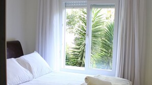 Ein Hotelzimmer mit Bett, weißen Vorhängen, einem Fenster mit Blick ins Grüne und einer braunen Tagesdecke.
