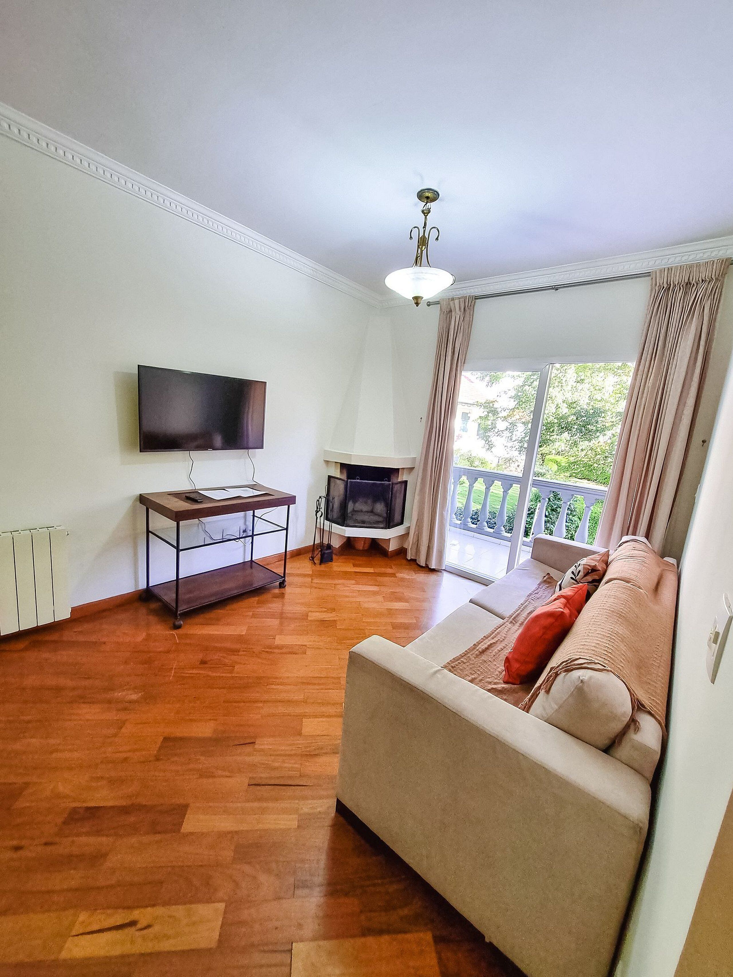 Uma sala de estar com televisão de tela plana, piso de madeira, sofá com almofadas, lareira e varanda com guarda-corpo.