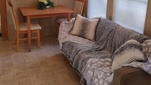 Un salón acogedor con una mesa de comedor de madera, dos sillas, un sofá con cojines estampados y un ventanal con persianas.
