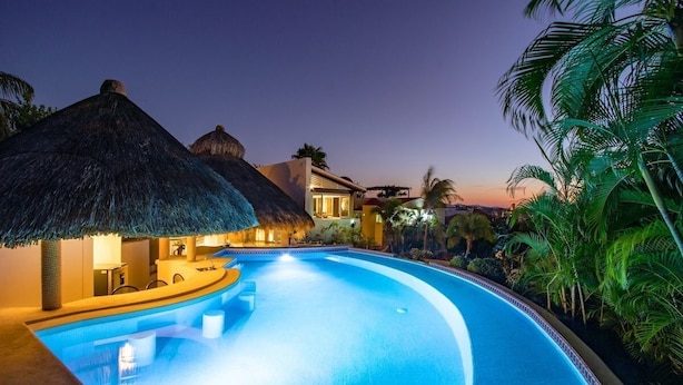 Una piscina de borde curvo, rodeada de palmeras y construcciones con techo de paja.