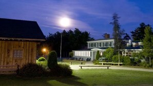 Una cabaña de madera y un edificio blanco con porche, rodeados de vegetación y un césped bien iluminado.