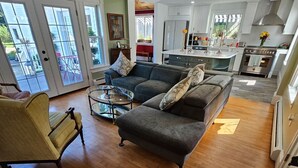 A spacious living area with a grey sofa, a glass coffee table, and a yellow armchair. A kitchen with white cabinets and a stainless steel stove is visible in the background. - Renovated Turn Of The Century Three Bedroom Farmhouse (Rockport)