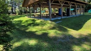 A covered picnic area with benches and tables, surrounded by a grassy field and tall trees. - Private Cabin with access to Rust Pond Ferris (Wolfeboro)