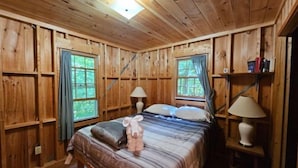 Un dormitorio de cabaña de madera con una cama, dos ventanas, una mesita de noche y una estantería con libros.