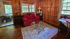 Interior de una cabaña de madera con un sofá rojo, una mesa de centro de vidrio y una zona de comedor con mesa y sillas.