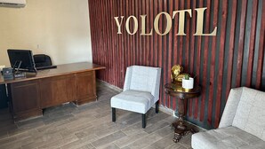 A reception area with a wooden desk, a computer, and two chairs. The wall has a vertical wooden panel with the text "YOLOTL" in gold letters. - Yolotl Sleep Experience (Piedras Negras)