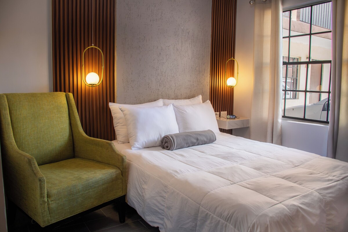 A hotel room with a bed, a green armchair, two wall-mounted lamps, and a window with a view of the outside.