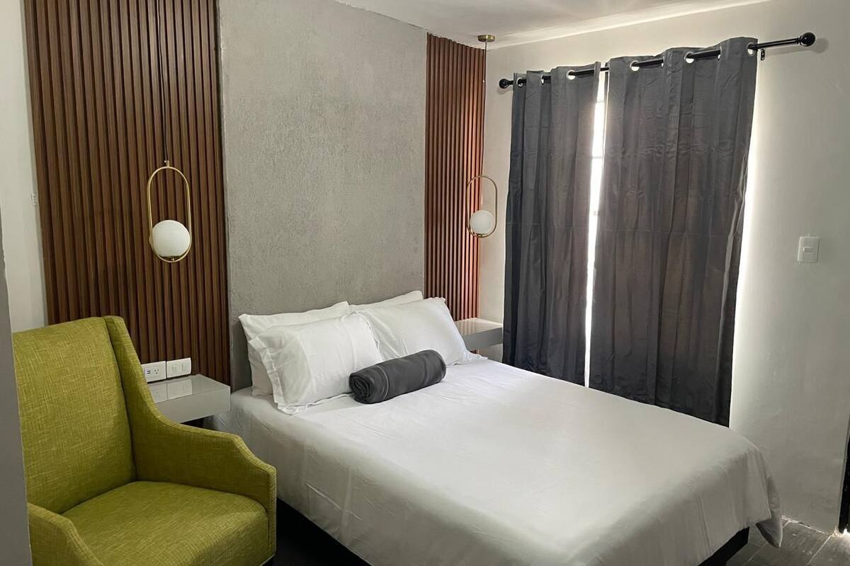 A neatly arranged hotel room with a single bed, a green armchair, and a bedside table with a lamp.