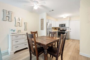 A dining area with a wooden table and chairs, a white dresser, and a kitchen in the background.