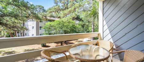 A balcony with wicker furniture, a glass table, and a view of trees and buildings.