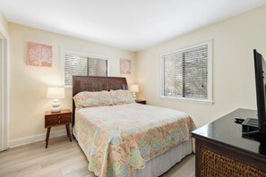 A bedroom with a bed, two bedside tables with lamps, a television on a stand, and two windows with blinds.