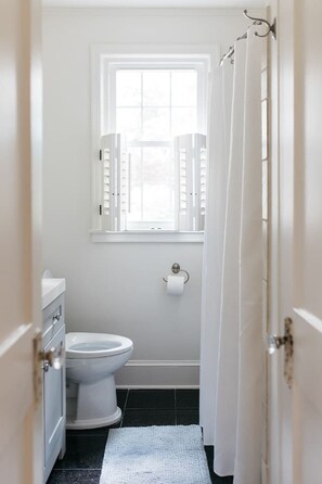 A small, white bathroom with a toilet, a window with open shutters, and a white shower curtain. - Walk to Marietta Square -The Cottage on Maple (Marietta)
